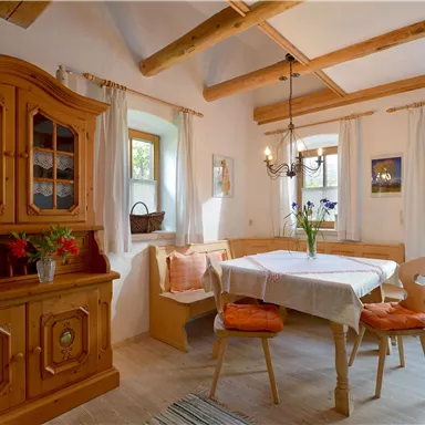 Ein gemütliches Esszimmer mit einem Holztisch und Stühlen. Es gibt eine Vitrine aus Holz und viel Tageslicht durch die Fenster.