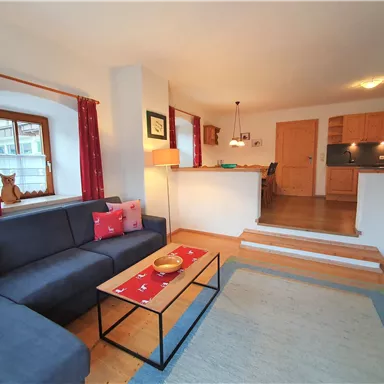 Ein gemütliches Wohnzimmer mit einem blauen Sofa, einem Holzkaffee Tisch und einer warmen Beleuchtung. Die offene Küche im Hintergrund ergänzt den einladenden Raum.