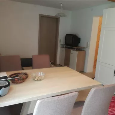 A bright living room with a dining table and chairs. In the background, a sofa and a kitchenette can be seen.