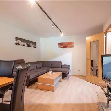 Ein modernes Wohnzimmer mit einem gemütlichen Sofa und einem Esstisch. Im Hintergrund ist ein Fernseher und ein Fenster mit Blick auf die Berge.