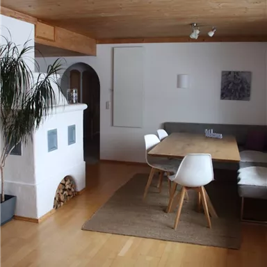 Ein modernes Wohnzimmer mit einem großen Holztisch und weißen Stühlen. Im Hintergrund ist ein Kamin und eine Pflanzenkübel zu sehen.