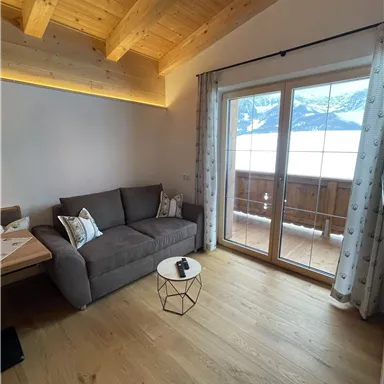 Ein gemütliches Wohnzimmer mit einem grauen Sofa und einem kleinen Tisch. Durch große Fenster sieht man einen Blick auf die Berge und einen Balkon.