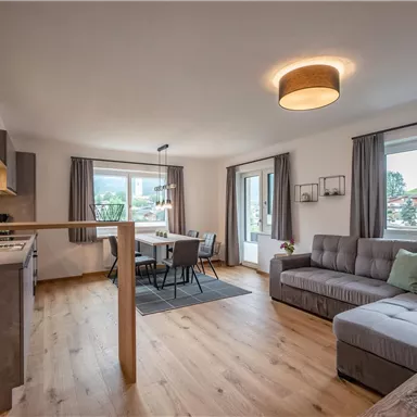 A modern and bright living room with a cozy couch and a dining table. Large windows let in plenty of daylight and offer a view outside.