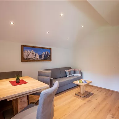 Ein gemütlicher Wohnraum mit Holzfußboden, einem Esstisch und einer modernen Couch. An der Wand hängt ein Fernseher und ein Bild mit Bergmotiven.