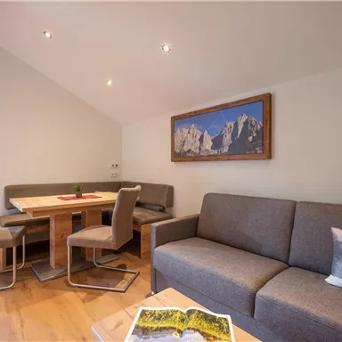 Ein modernes Wohnzimmer mit einem grauen Sofa und einem Esstisch. An der Wand hängt ein Landschaftsbild und es gibt Fenster mit Vorhängen.