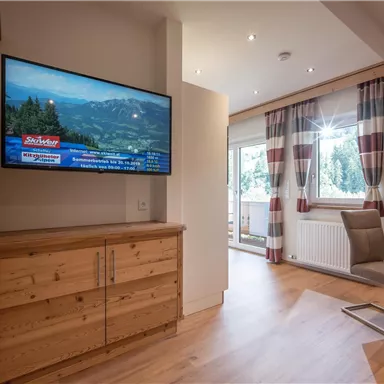 Ein modernes Wohnzimmer mit Holzoberflächen und einem großen Fernseher. Helle Vorhänge und ein Esstisch schaffen eine einladende Atmosphäre.