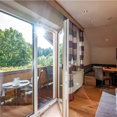 Ein heller Wohnraum mit großen Fenstern und Blick auf grüne Bäume. Im Vordergrund sind ein Tisch und Stühle sichtbar.