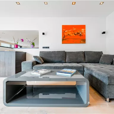 Ein modernes Wohnzimmer mit einem grauen Sofa und einem stylischen Tisch. An der Wand hängt ein auffälliges Kunstwerk in Orange.
