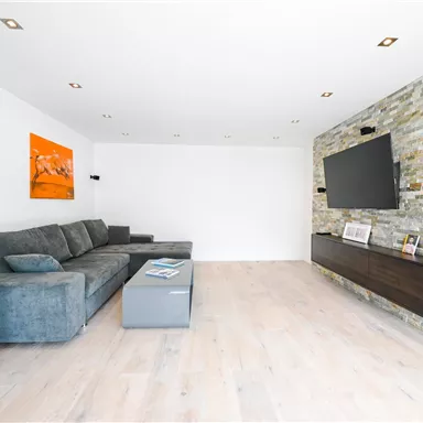 Ein modernes Wohnzimmer mit einem grauen Sofa und einem Fernseher an der Wand. Die Wand ist aus Stein und es gibt einen niedrigen Tisch in der Mitte des Raumes.