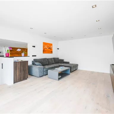 Ein modernes Wohnzimmer mit einem grauen Sofa und einem Fernseher an der Wand. Der Raum hat helle Wände und einen eleganten, minimalistischen Stil.