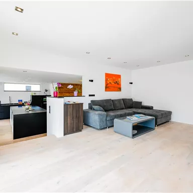 Ein modernes Wohnzimmer mit einem grauen Sofa und hellem Holzfußboden. An der Wand hängt ein orangefarbenes Bild, und der Raum wirkt hell und einladend.