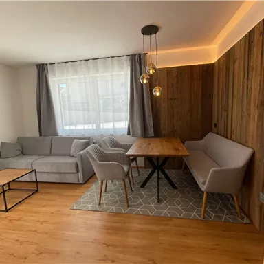 A modern living room with a gray sofa, a wooden table, and light walls. The large window front lets in plenty of light and creates a friendly atmosphere.