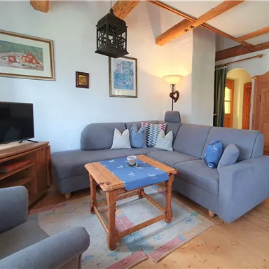 Ein gemütliches Wohnzimmer mit einer grauen Couch und einem Holztisch. An der Wand hängen Bilder, und im Hintergrund ist ein Fernseher zu sehen.