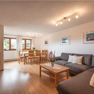 Ein gemütliches Wohnzimmer mit einer grauen Couch und einem Holztisch. Im Hintergrund befindet sich ein Essbereich mit einem Esstisch und Stühlen.