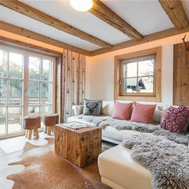 Ein gemütliches Wohnzimmer mit Holzbalken und rustikalen Möbeln. Große Fenster lassen viel Licht herein und bieten Blick auf die Natur.