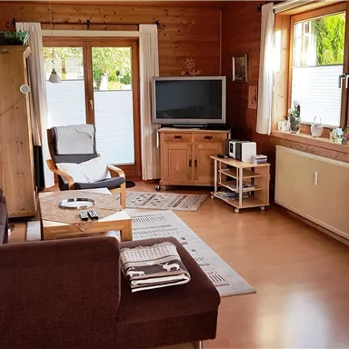 A cozy living space with a sofa, an armchair, and a television. Large windows let in plenty of daylight and offer a view of the outdoors.