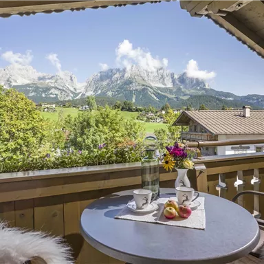 A beautiful balcony with a view of majestic mountains and a green landscape. On the table are two cups of coffee and a bouquet of flowers.