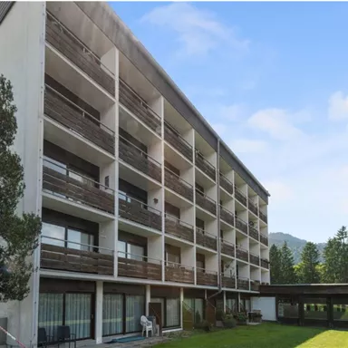 A modern multi-story building with balconies and large windows. In the foreground, there is a green lawn and a covered area.