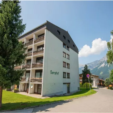 A modern building with several floors and balconies, surrounded by green trees. In the background, mountains and a clear blue sky can be seen.