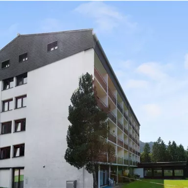 A modern building with several floors and large windows. Surrounded by a green meadow and trees.