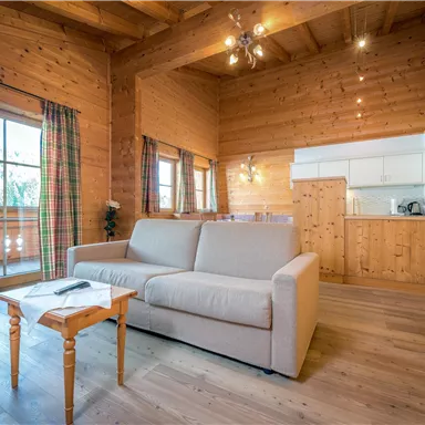 Ein gemütlicher Wohnraum mit Holzverkleidung und großen Fenstern. Im Vordergrund steht ein helles Sofa und ein kleiner Tisch.