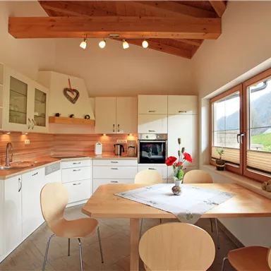 Eine moderne Küchenzeile mit weißen Schränken und Holzdetails. Ein runder Tisch mit Stühlen steht vor großen Fenstern, die einen Blick auf die grüne Landschaft bieten.