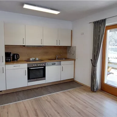 Eine moderne Küche mit einem Herd, einem Kühlschrank und einer Sitzecke. Große Fenster sorgen für viel Tageslicht und Blick auf die Schneelandschaft draußen.