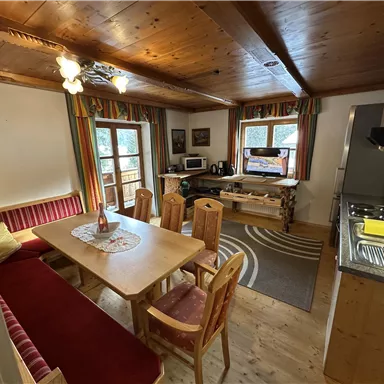 Ein gemütlicher Wohnraum mit einem großen Esstisch, Stühlen und einer Küche. Das Zimmer hat Holzdecken und große Fenster, die viel Licht hereinlassen.