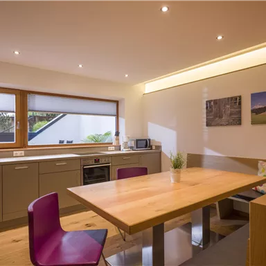 Eine moderne Küche mit einem großen Holztisch und pinken Stühlen. Die Wände sind hell und es gibt einige Bilder.