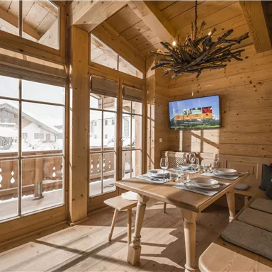 Eine gemütliche Stube aus Holz mit großen Fenstern und einem Esstisch. An der Wand hängt ein Fernseher, und der Blick geht auf die schneebedeckte Landschaft draußen.