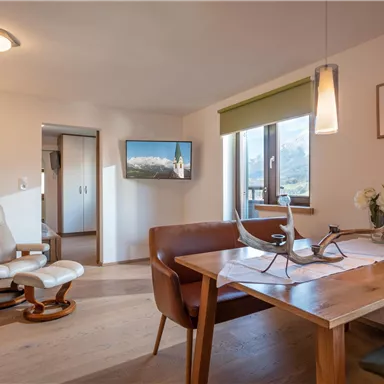 Ein gemütlicher Wohnbereich mit einem Esstisch, einem bequemen Stuhl und Wänden in neutralen Farben. Durch das Fenster ist ein schöner Ausblick zu sehen, während ein Fernseher an der Wand hängt.