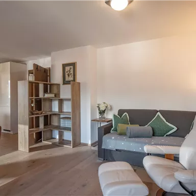 Ein gemütliches Wohnzimmer mit einem grauen Sofa, einer weißen Leselampe und einem Bücherregal. Der Raum hat helle Wände und einen modernen, einladenden Look.