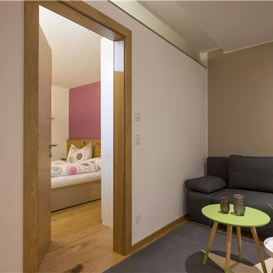 Ein gemütlicher Raum mit einem grauen Sofa und einem kleinen Tisch. Im Hintergrund ist ein Schlafzimmer mit einem brassfarbigen Bett zu sehen.