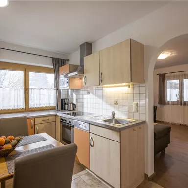 A bright kitchen with modern furnishings and a dining table. In the background, a cozy living room is visible.