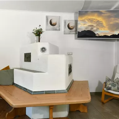 Ein gemütlicher Wohnraum mit einem weißen Kachelofen und einem großen Fernseher an der Wand. Eine Holzbank und ein moderner Stuhl laden zum Entspannen ein.