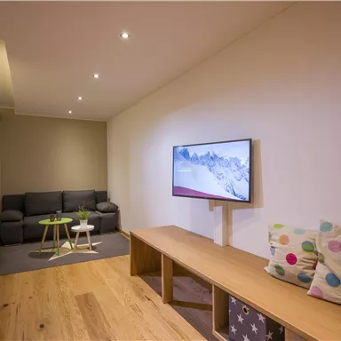 Ein modernes Wohnzimmer mit einem grauen Sofa und einem Fernseher an der Wand. Der Holztisch und der Boden verleihen dem Raum eine helle, gemütliche Atmosphäre.