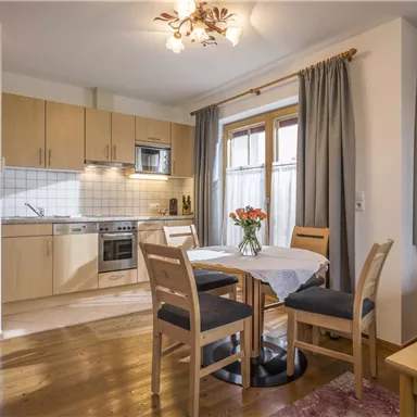 Eine moderne Küche mit Essbereich und einem Tisch für vier Personen. Die Fenster lassen viel Tageslicht herein und die Umgebung wirkt einladend.