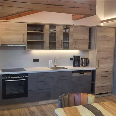 Eine moderne Küche mit Holzschränken und einem klaren Design. Im Vordergrund befindet sich ein Esstisch mit mehreren bunten Stühlen.