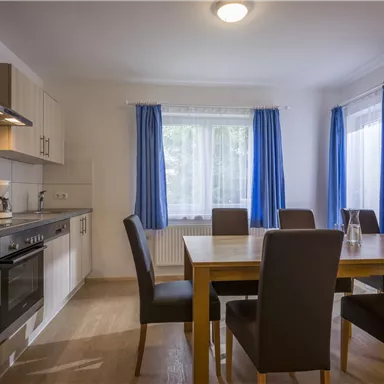 Eine helle Küche mit einem großen Esstisch und Stühlen. Vor den Fenstern hängen blaue Vorhänge und es gibt moderne Küchengeräte.