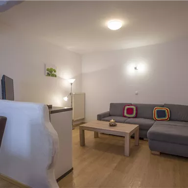 Ein modernes Wohnzimmer mit einem grauen Sofa und einem Holztisch. Die Wände sind hell und die Möbel sorgen für eine gemütliche Atmosphäre.