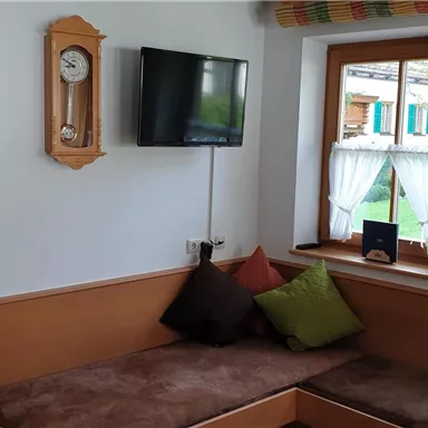 A cozy room with seating and colorful cushions. A clock and a television hang on the wall, and the window offers a view outside.