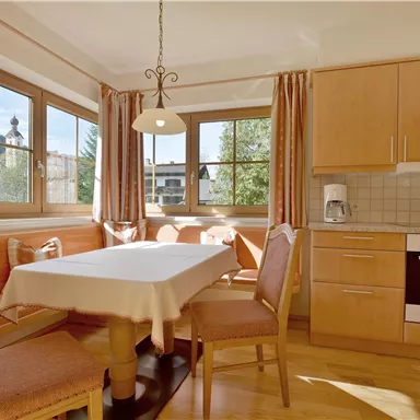 Eine helle Küche mit einem Essbereich und großen Fenstern. Der Tisch ist mit einem weißen Tischtuch gedeckt und die Wände sind in warmen Farbtönen gehalten.