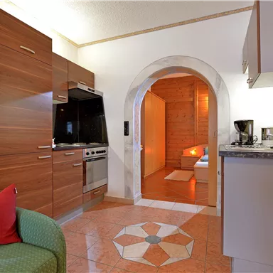 A modern kitchen with wooden cabinets and an open living area. In the background, a cozy room is visible, accessible through an arch.