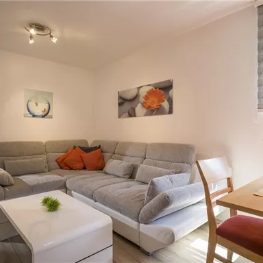 A cozy living room with a gray couch and a white table. There are some plants and pictures on the walls.