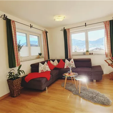 Ein gemütliches Wohnzimmer mit einem grauen Sofa und bunten Kissen. Große Fenster bieten einen schönen Blick auf die Berge und viel Tageslicht.