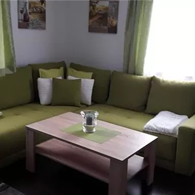 Eine gemütliche Wohnzimmerecke mit einem grünen Sofa und Kissen. Ein Holztisch steht in der Mitte, umgeben von hellen Vorhängen und Bildern an der Wand.