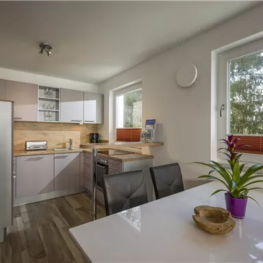 A modern kitchen with bright cabinets and a dining table. Large windows provide a view of trees outside.
