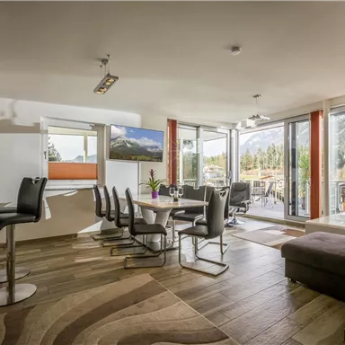 A modern living room with large windows and plenty of natural light. The space features a dining area and stylish furniture.