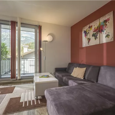 A modern living room with a gray sofa and a white table.  
Large windows offer a view of the mountains and the balcony.
