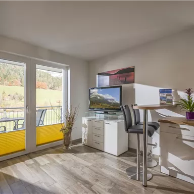 A modern kitchen with a dining table and chairs. Large windows provide a view of the green landscape outside.
